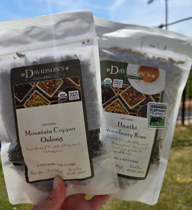 Two bags of Davidson's Organics loose-leaf tea held in a hand outdoors. The left bag is Mountain Copper Oolong, and the right bag is Haathi Strawberry Rose.