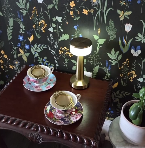 A styled interior scene showing a small illuminated table lamp on a dark wooden side table, alongside two floral teacups and saucers, set against a dark floral wallpaper. A potted succulent is visible on the right.