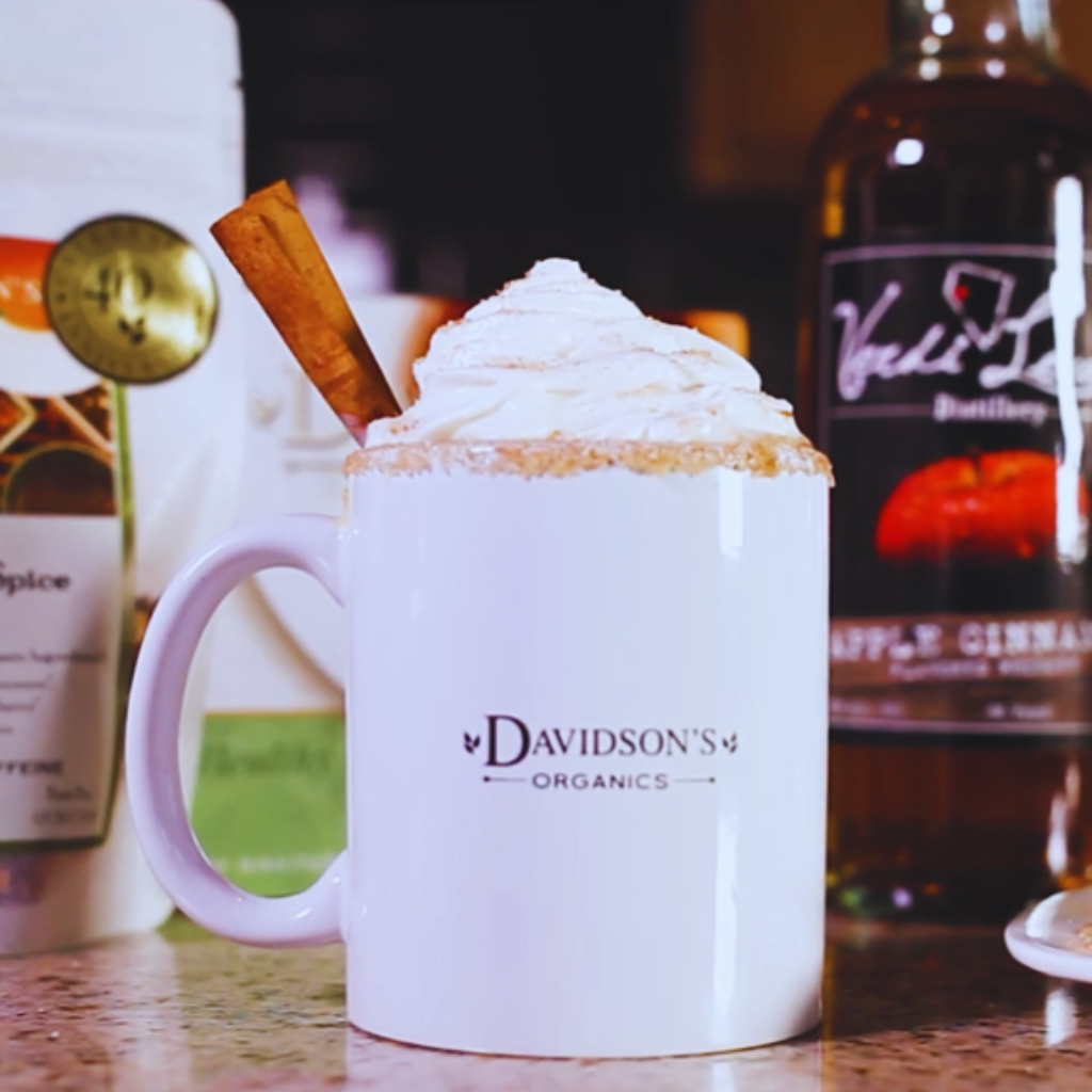 White mug with whipped cream and a cinnamon stick, surrounded by a bottle of伏特加 and a box of Davidson's Organics spice mix.