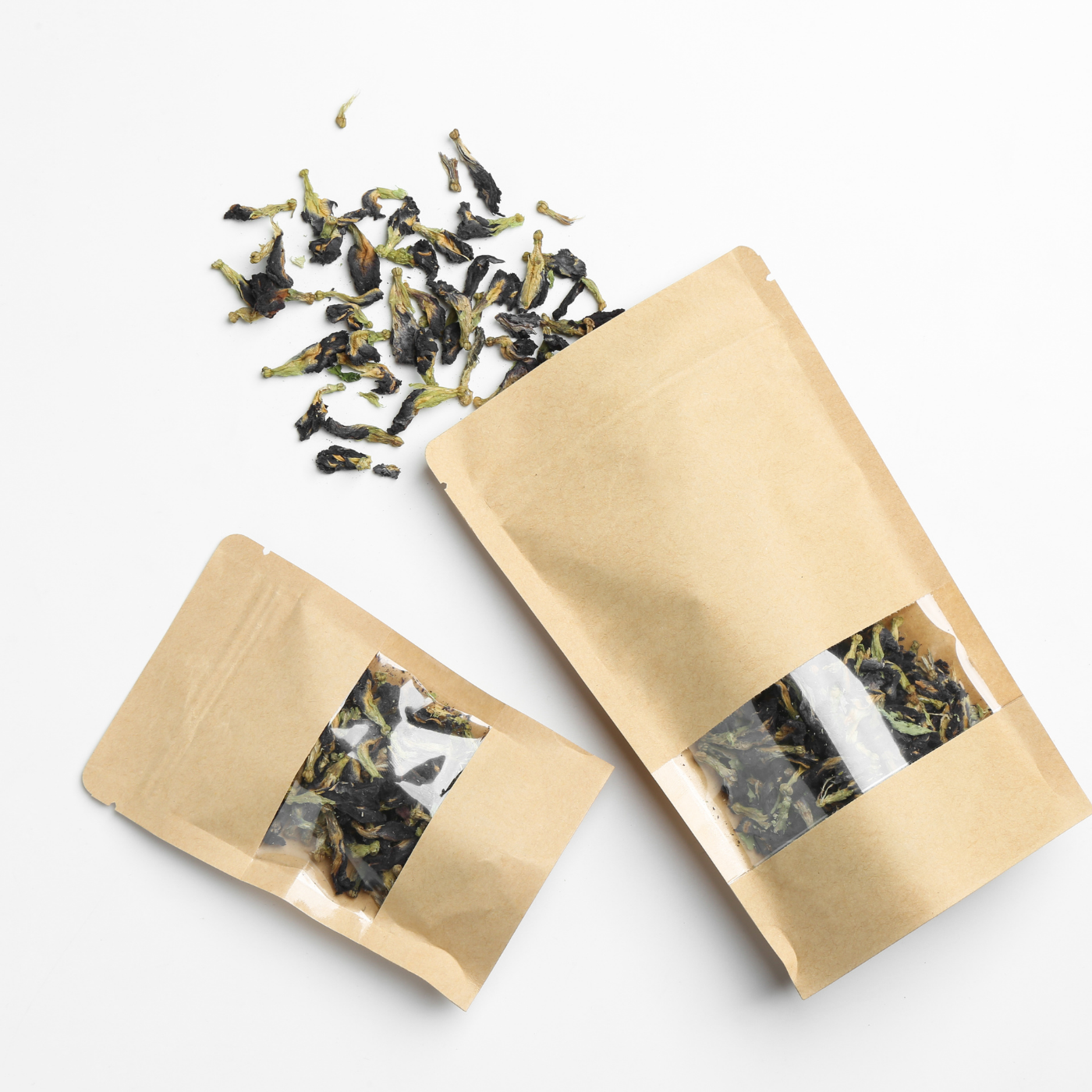 Two small kraft paper stand-up pouches with clear windows displaying loose dried tea leaves, with a few tea leaves scattered above the bags on a white background.