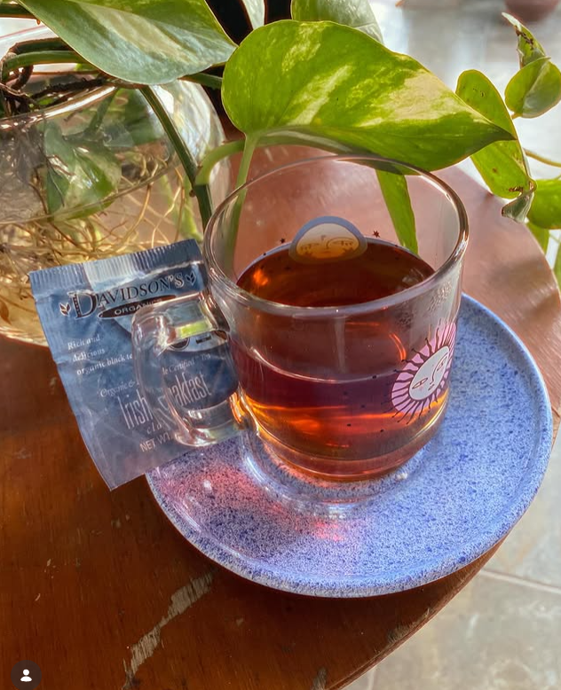A clear glass mug filled with amber tea sits on a speckled blue saucer on a wooden table, with a tea packet beside it and green plant leaves hanging over the cup.