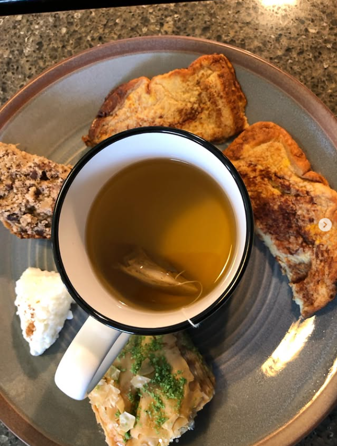 A top-down view of a breakfast spread on a round gray plate, featuring a white mug filled with tea, toast, a cracker, and a piece of salmon with green herbs, set on a speckled granite countertop.