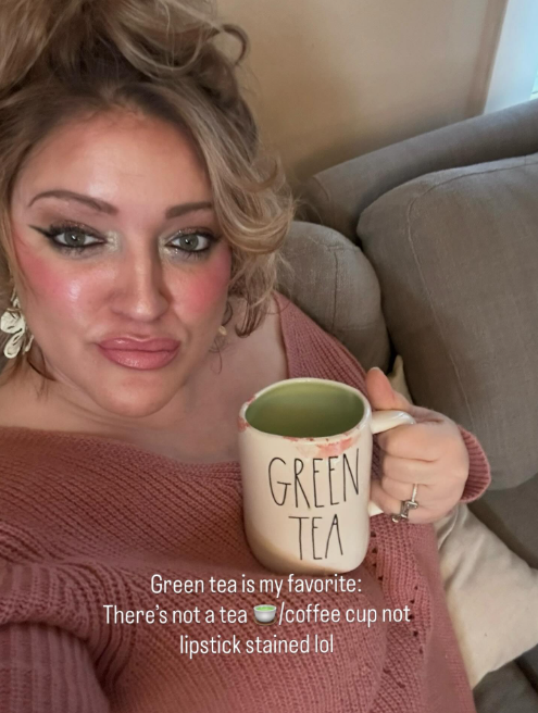 A woman in a rose knit sweater holds a ceramic mug with 'GREEN TEA' printed on it, sitting on a beige couch. Text overlay reads: 'Green tea is my favorite: There's not a tea cup/coffee cup not lipstick stained lol'.