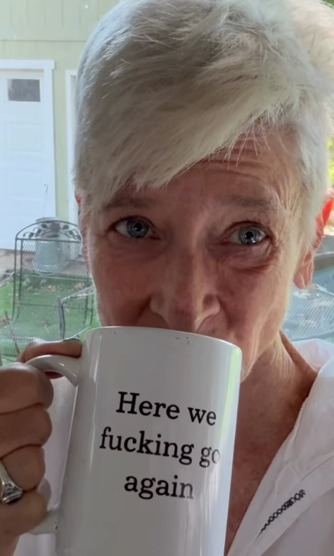 A close-up of a person with short white hair holding a white ceramic mug with the printed text 'Here we fucking go again'.