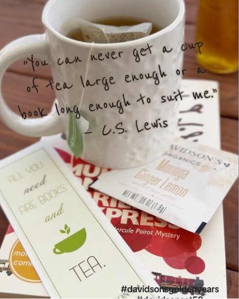 A white ceramic mug filled with tea and a tea bag, placed on top of several tea and book-themed cards and packets. A handwritten quote is overlaid across the mug: “You can never get a cup of tea large enough or a book long enough to suit me.