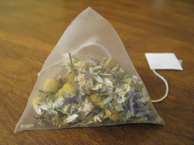 A triangular mesh tea bag filled with dried chamomile flowers and other loose herbal tea ingredients, sitting on a wooden surface with its white paper tag hanging to the right.