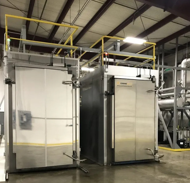 Two large industrial stainless steel machines in a warehouse setting, featuring yellow safety railings and metal support structures.