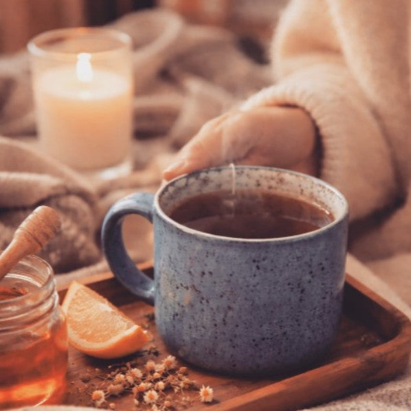 A steaming blue speckled ceramic mug filled with tea on a wooden tray, with a hand in a cream sweater holding the handle, alongside a honey jar, orange slice, and a lit candle in the background.