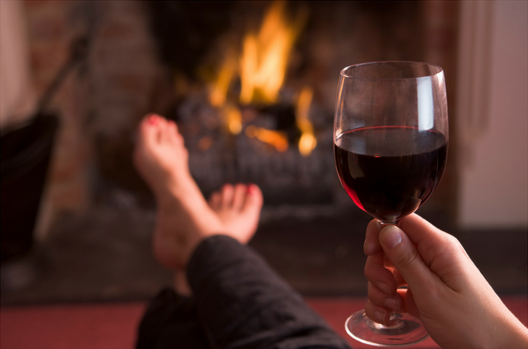 Person relaxing by a warm fireplace with a glass of red wine.