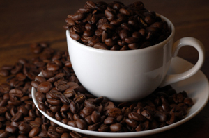 White cup overflowing with dark roasted coffee beans on a wooden surface.