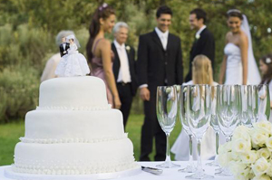 Tiered white wedding cake with a couple and guests standing nearby.